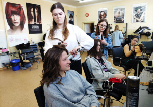 Dwie dziewczynki siedzą na fotelach fryzjerskich i są czesane przez przyszłe fryzjerki.